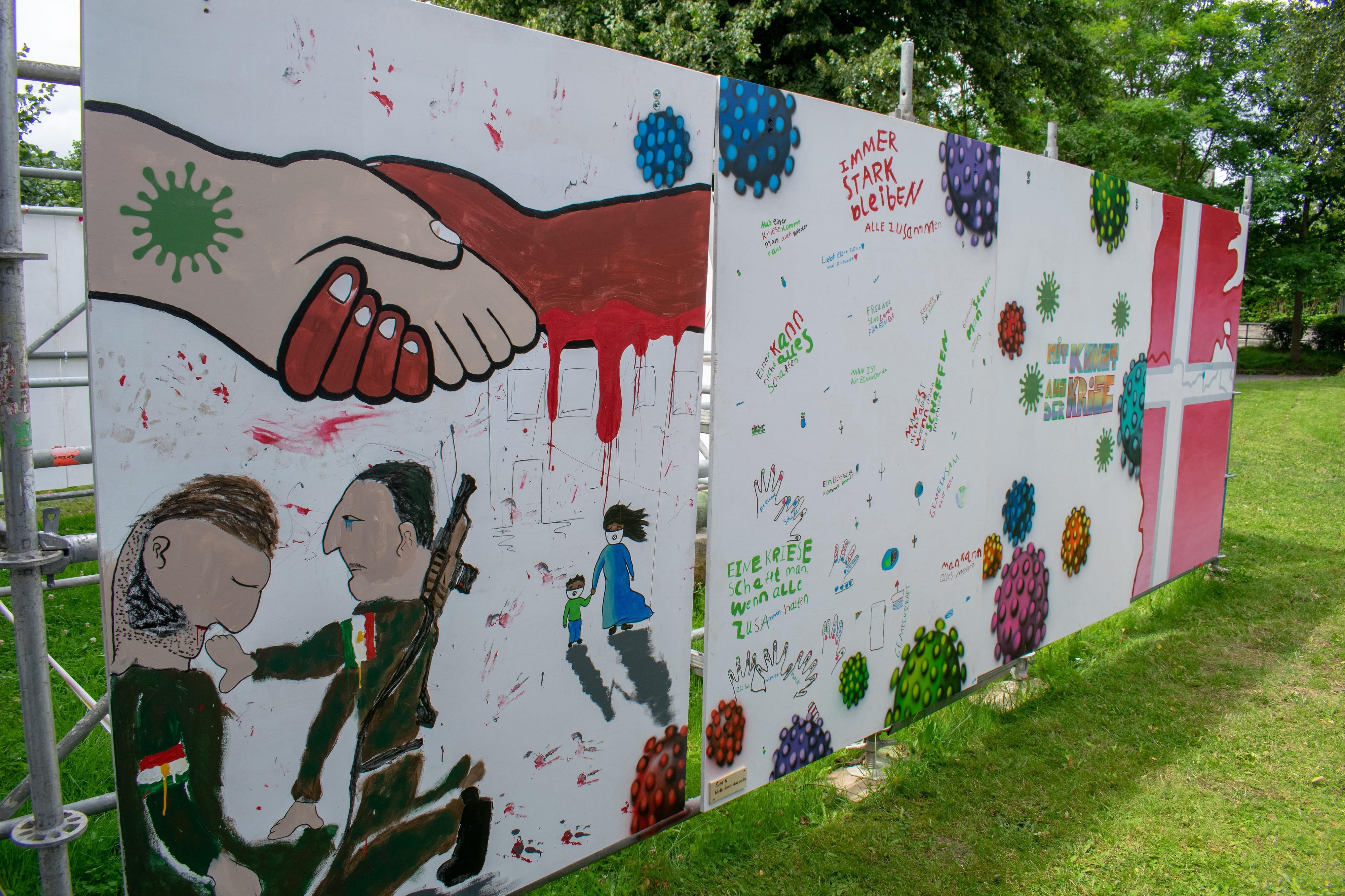 Ein Kunstwerk, das zwei Flüchtlinge mit Blick auf zwei Soldaten zeigt und Hände die das Corona-Virus und den Krieg symbolisieren
