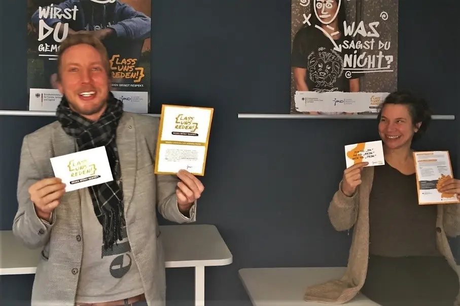 Das Foto zeigt zwei Respekt Coaches, die lachend Einladungen für die Veranstaltung "Lass uns reden" in die Kamera halten.