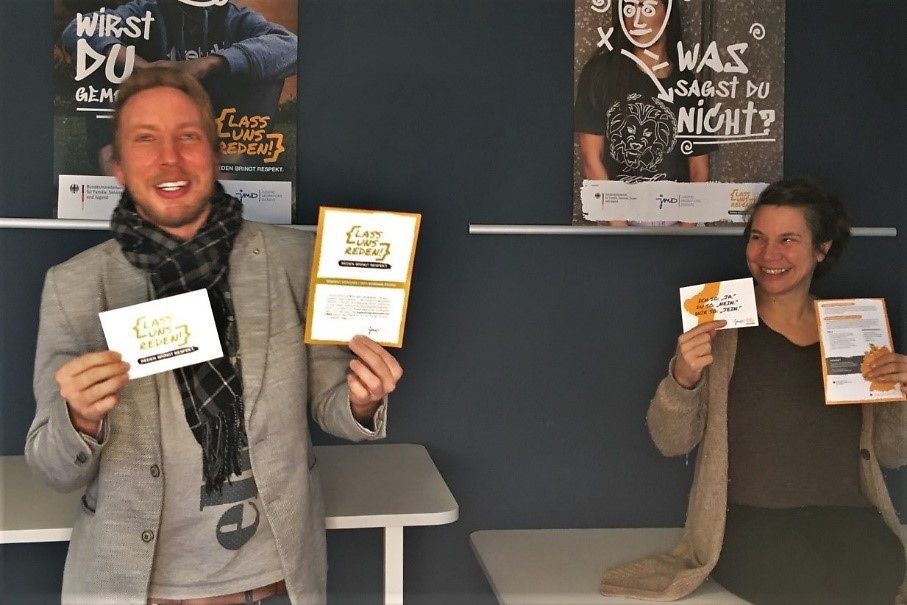 Das Foto zeigt zwei Respekt Coaches, die lachend Einladungen für die Veranstaltung "Lass uns reden" in die Kamera halten.