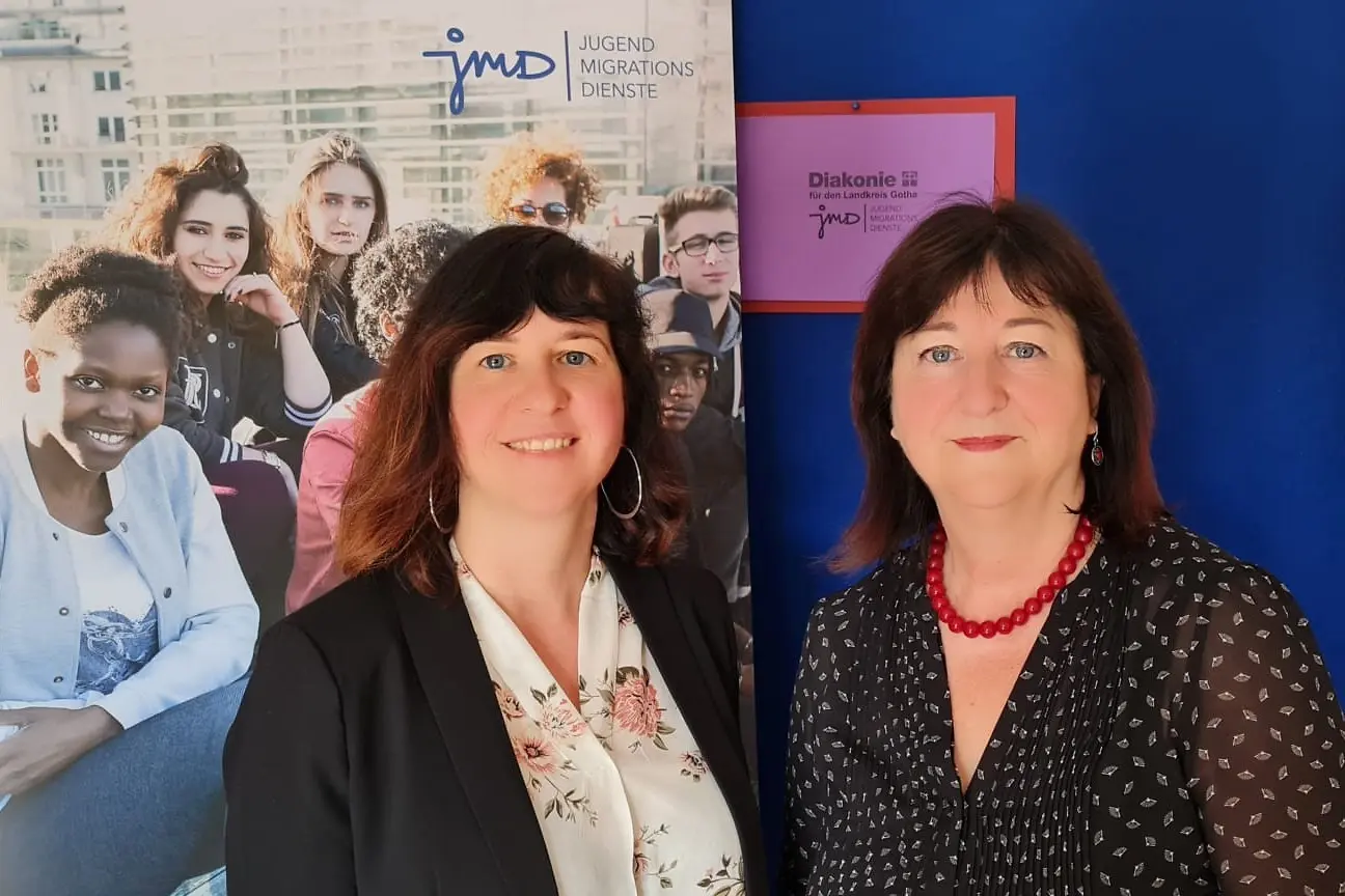 Frau Sigrid Ansorg und Ihre Tochter Kathrin Ansorg stehen vor einem Plakat des Jugendmigrationsdientes