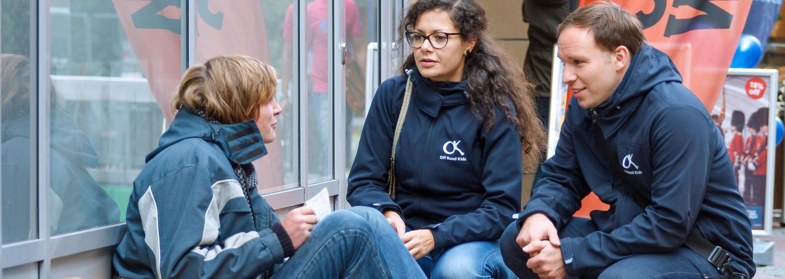 Eine junge Frau und ein junger Mann sprechen mit einer obdachlosen, jungen Person auf der Straße