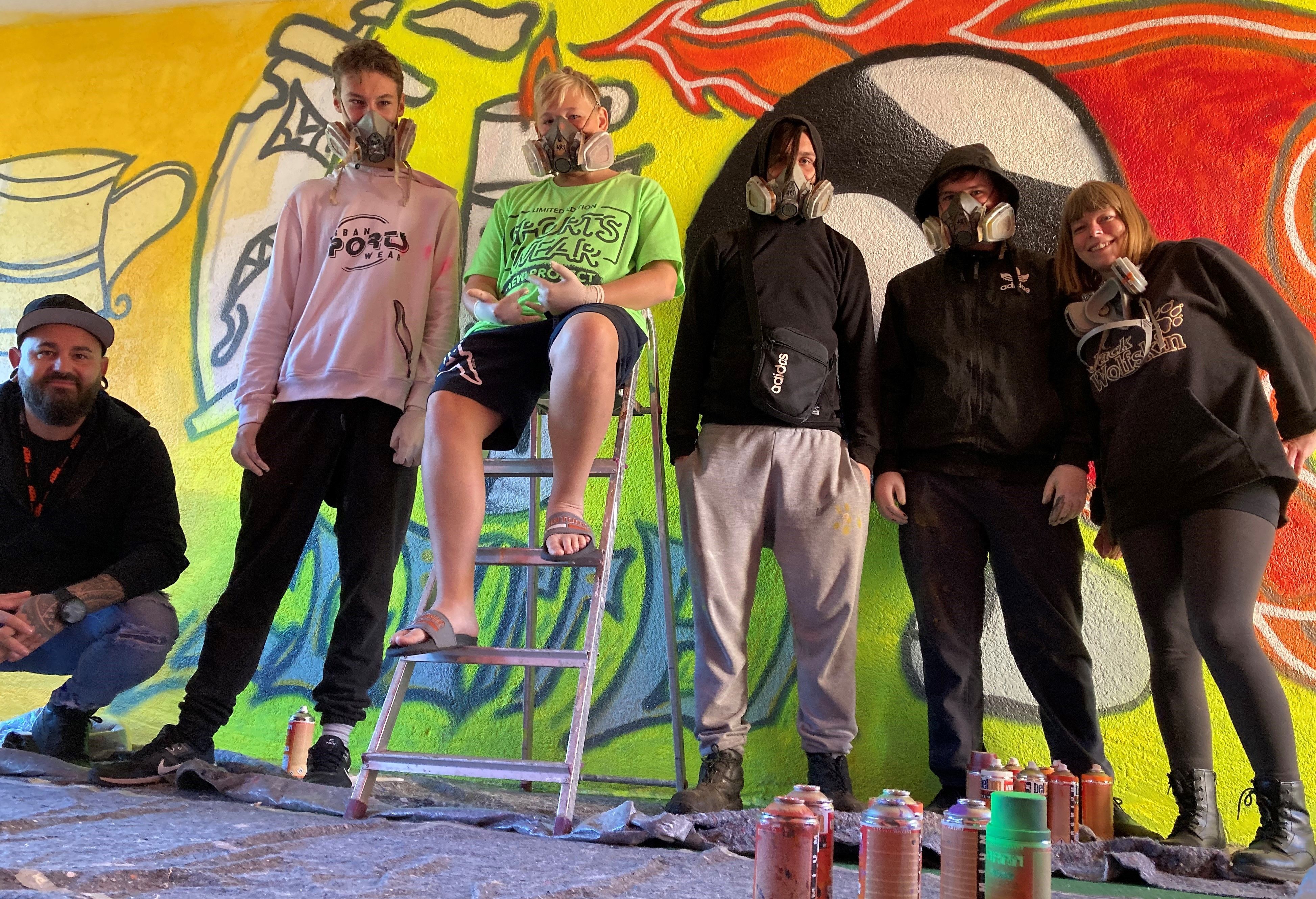 Sechs Jugendliche posieren vor einer Wand mit ihrem Graffiti Kunstwerk.