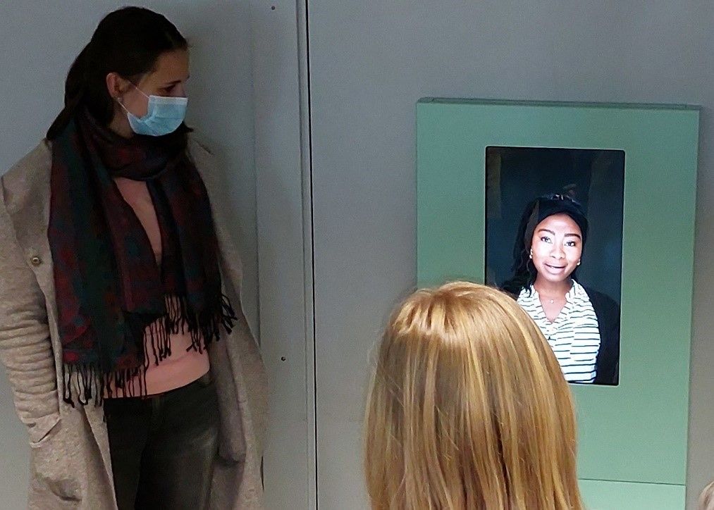 Zwei junge Frauen stehen vor einem Bildschirm und hören einem Video-Interwiews zu.