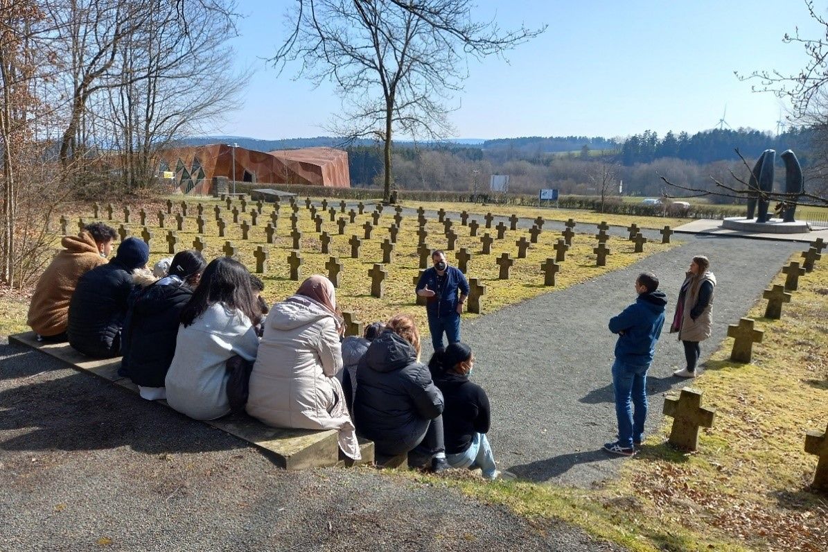 Viele Jugendliche sitzen auf einer Treppe vor Kreuzen in einem ehemaligen SS-Lager. 