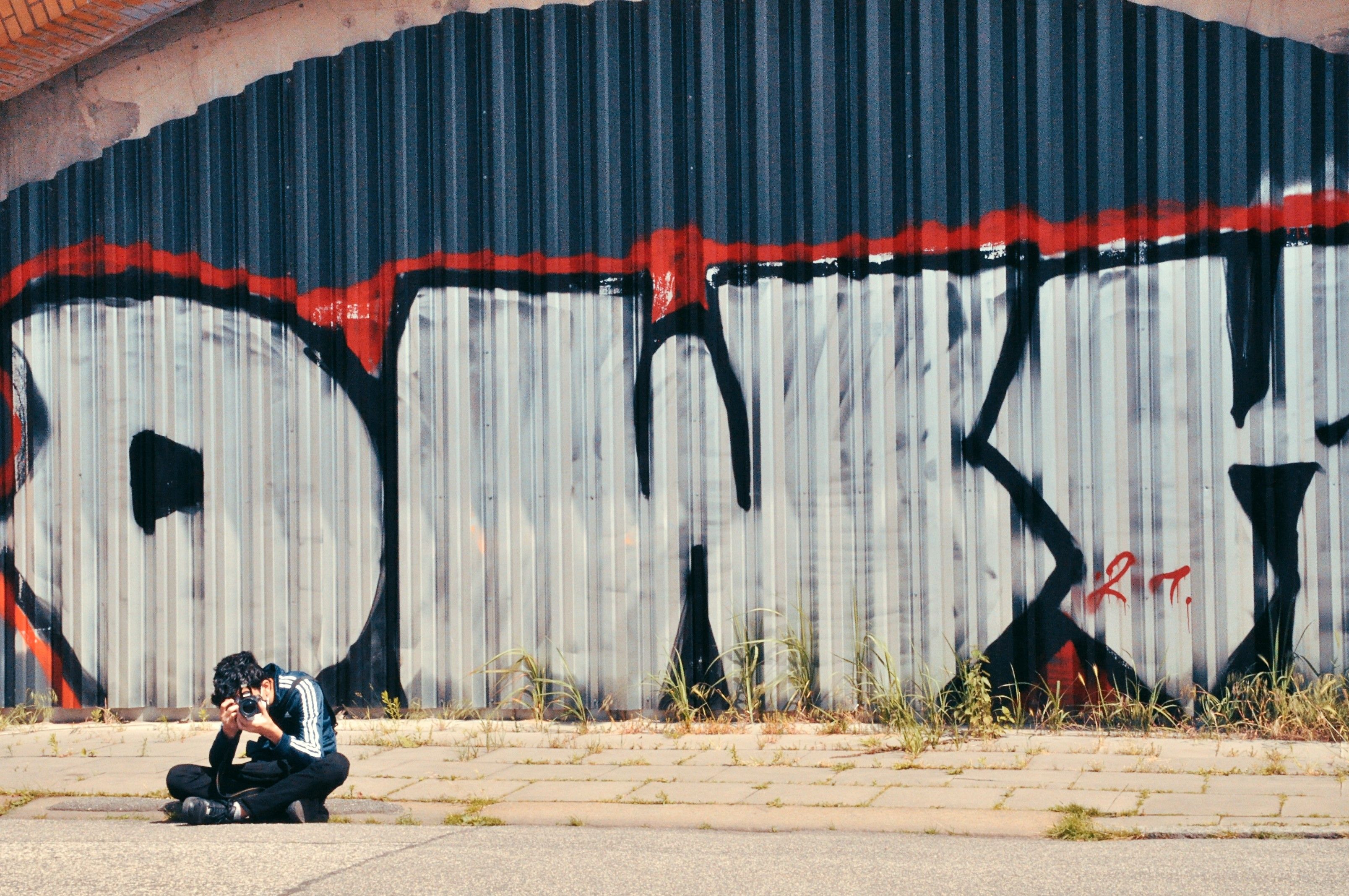 Das Foto zeigt eine fotografierende Person vor einer mit Graffiti bemalten Wand sitzend