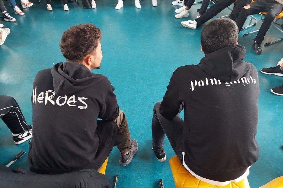 Zwei Männer sitzen mit dem Rücken zur Kamera. Einer von den beiden trägt einen Hoodie mit der Aufschrift Heroes. 