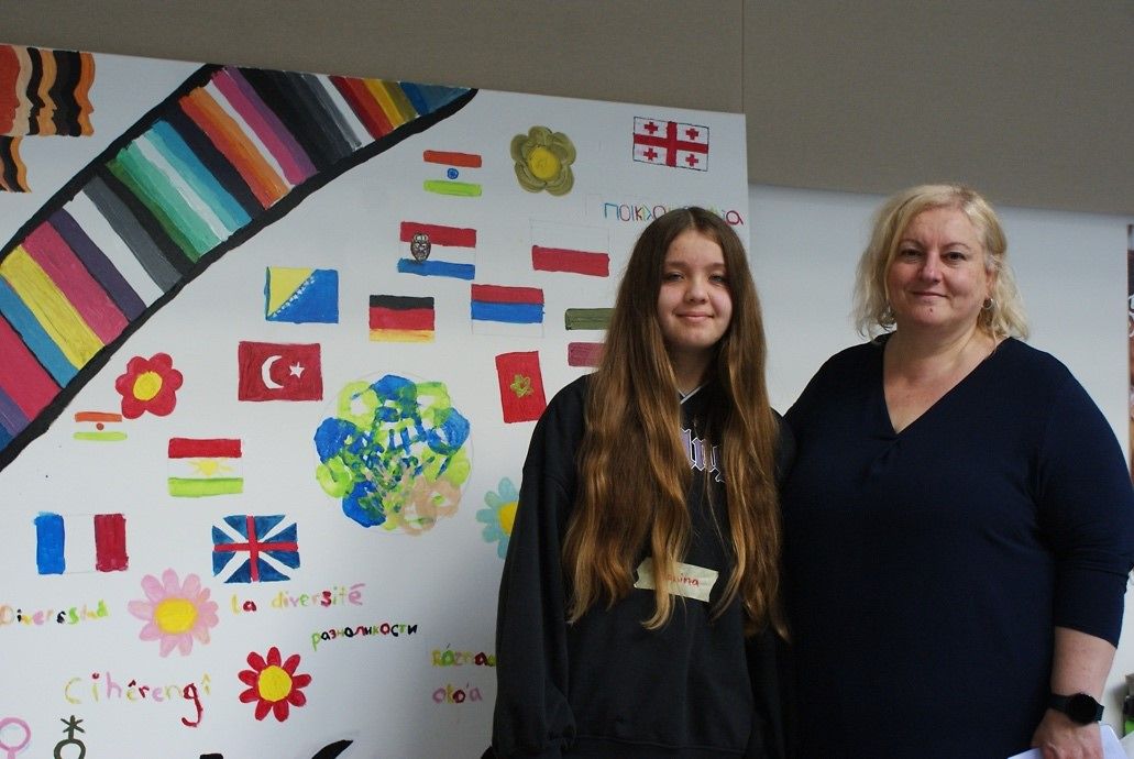 Eine Schülerin und Janina Pavlovic stehen vor einer Leinwand mit vielen verschiedenen Flaggen drauf