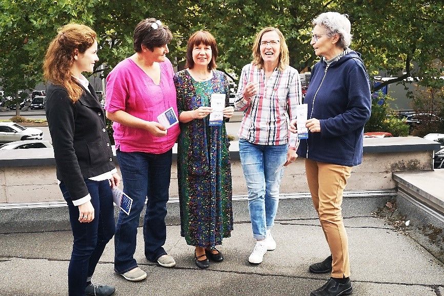 Fünf Frauen stehen in einem Halbkreis mit einem Flyer in der Hand.