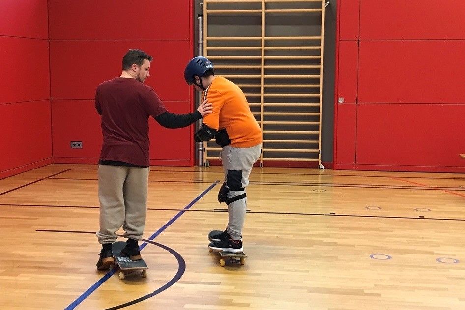 Ein Erwachsener auf einem Skateboard und ein Jugendlicher auf einem Skateboard mit Schutzausrüstung in einer Turnhalle.