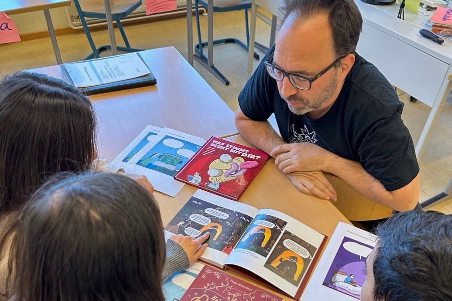 Herr Piero Masztalerz sitz mit drei Jugendlichen an einem Tisch und guckt sich Karikaturen an.