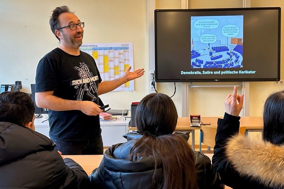 Herr Piero Masztalerz steht in einem Klassenraum vor einem Bildschirm und guckt sich mit den Schülern Karikaturen an.