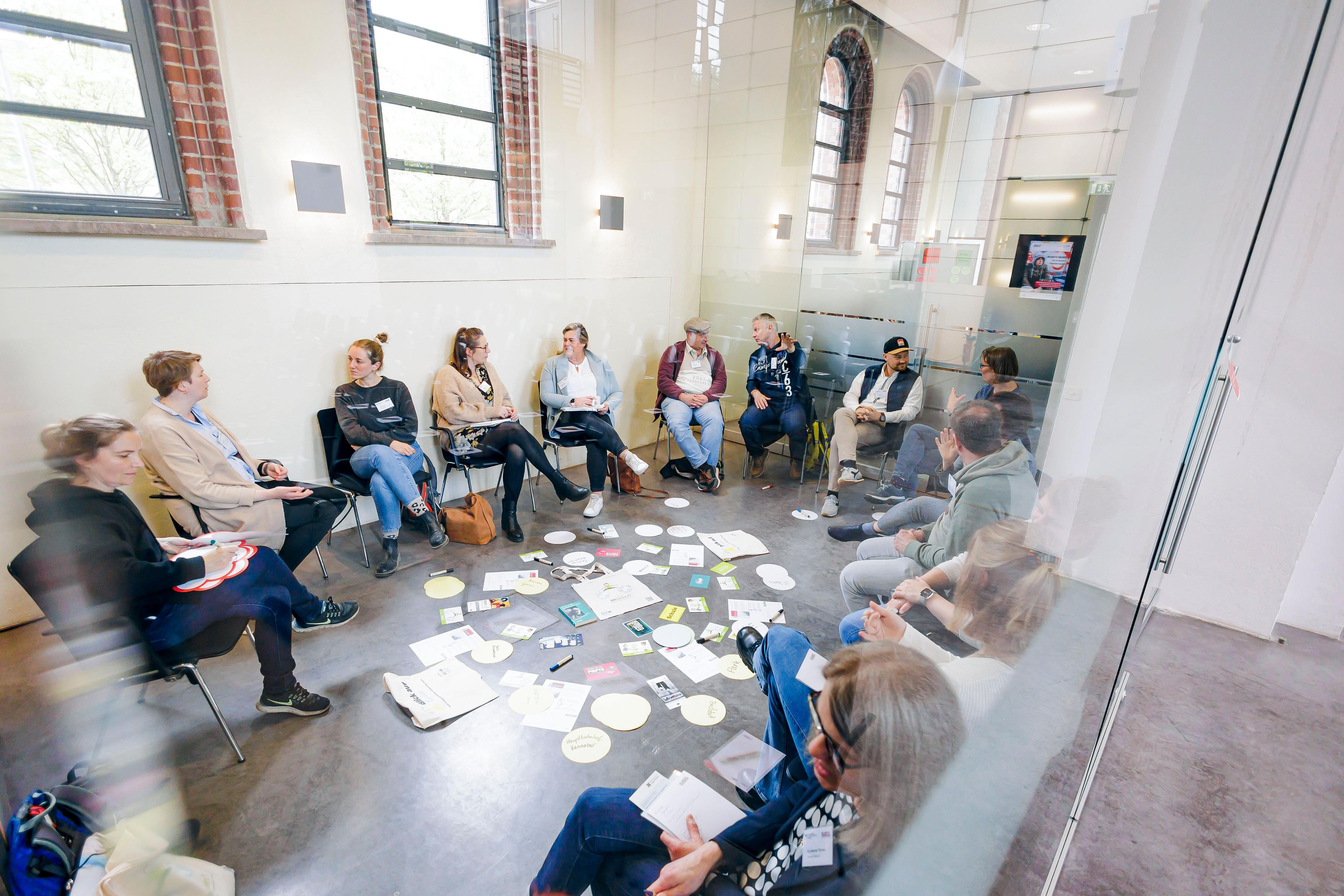 Eine Gruppe Erwachsener sitzt bei einem Workshop im Kreis und in der Mitte liegen viele zettel und Stifte