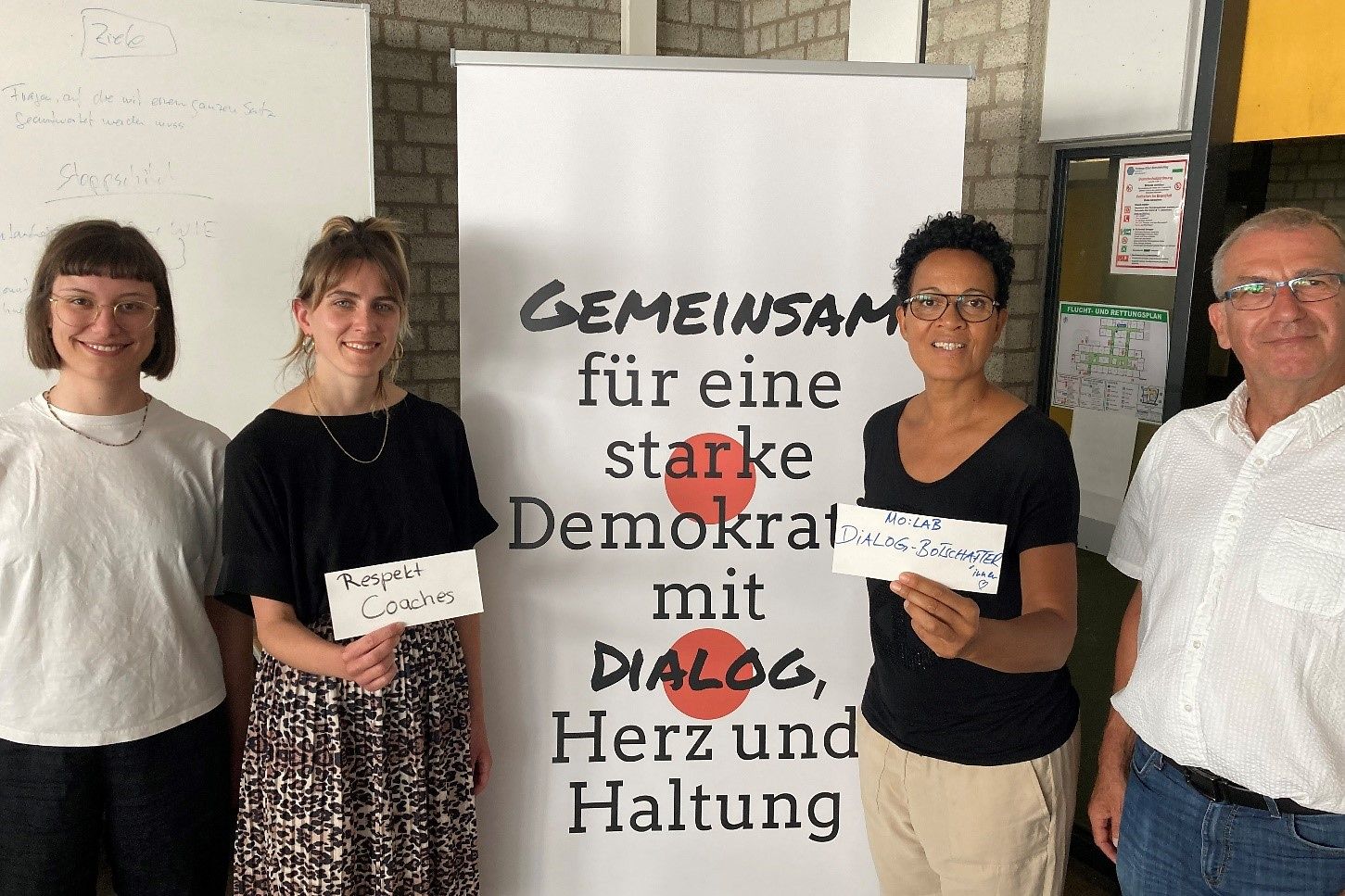 Drei Frau und eine Mann vor einem Banner mit der Aufschrift: Gemeinesam für eine starke Demokratie mit Dialog, Herz und Haltung