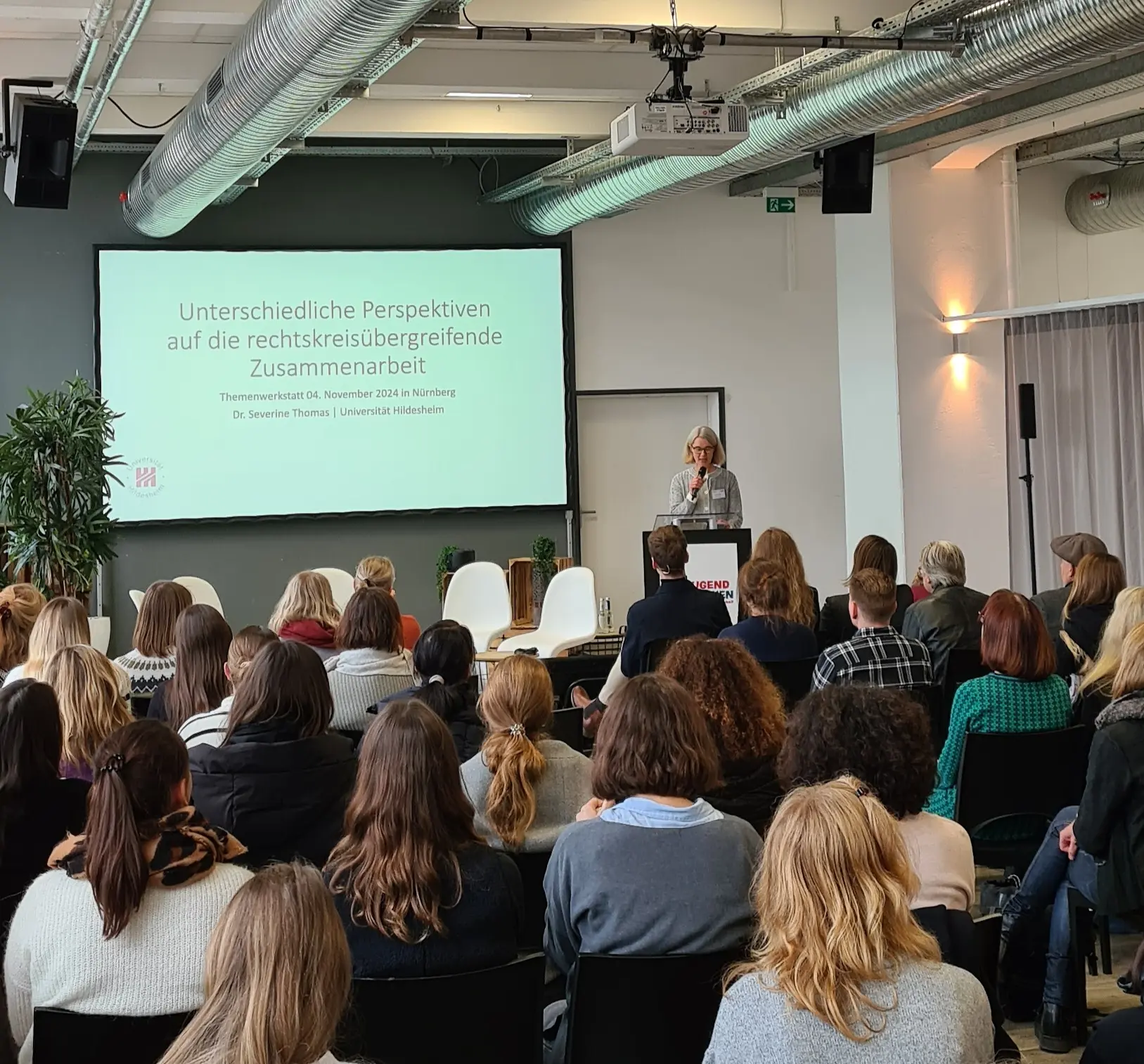 Eine Frau steht neben einer Leinwand und hält einen Vortrag vor einer großen Gruppe.