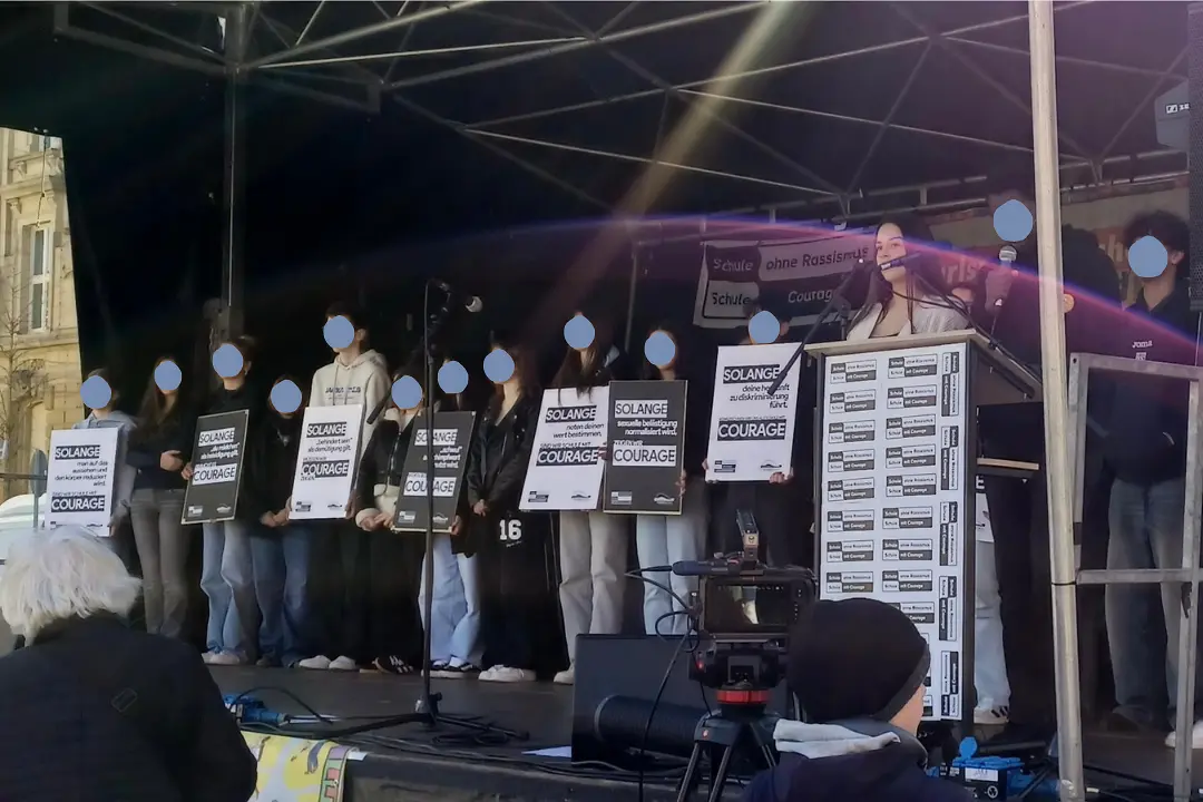Schüler*innen stehen mit Plakaten gegen Rassismus auf einer Bühne.