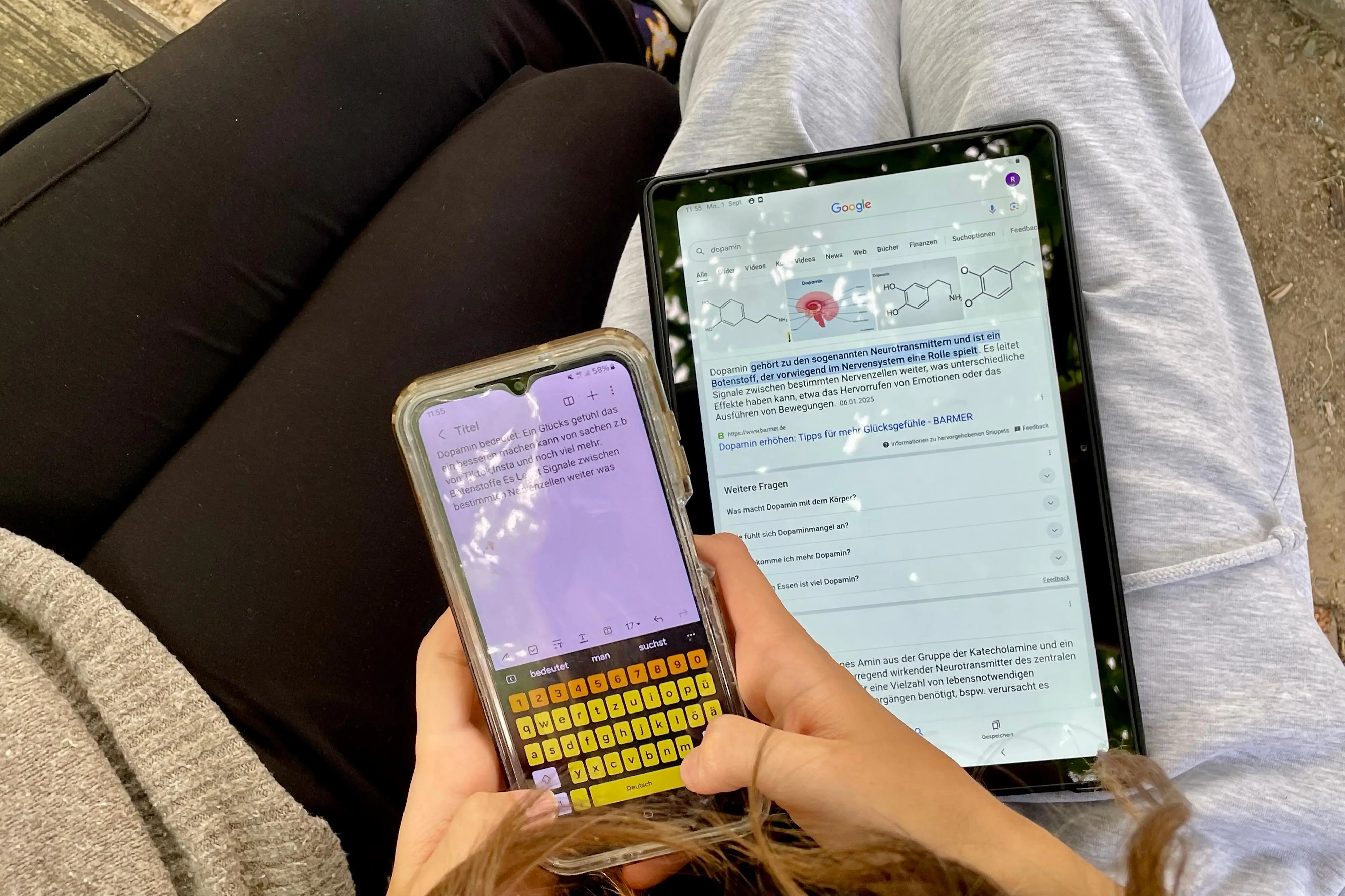 Ein junger Mensch hat ein Tablet auf dem Schoß und ein Handy in der Hand.