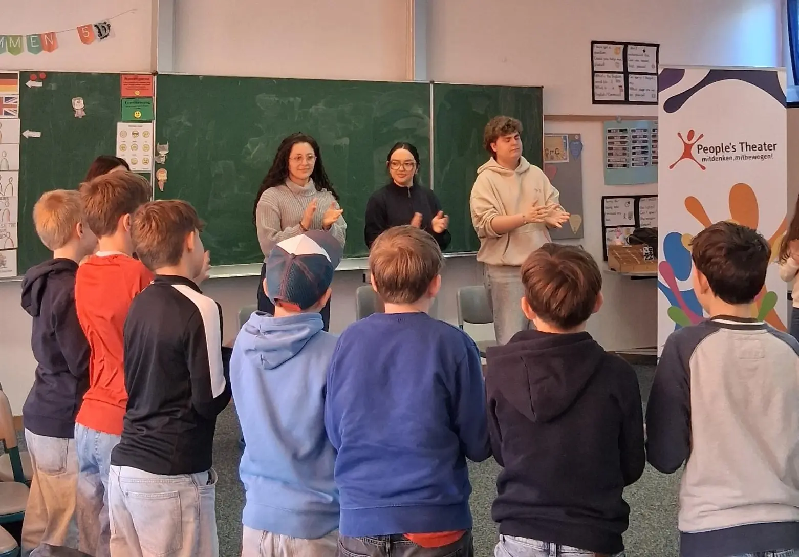 Eine Gruppe steht in einer Klasse vor einer Tafel im Kreis.